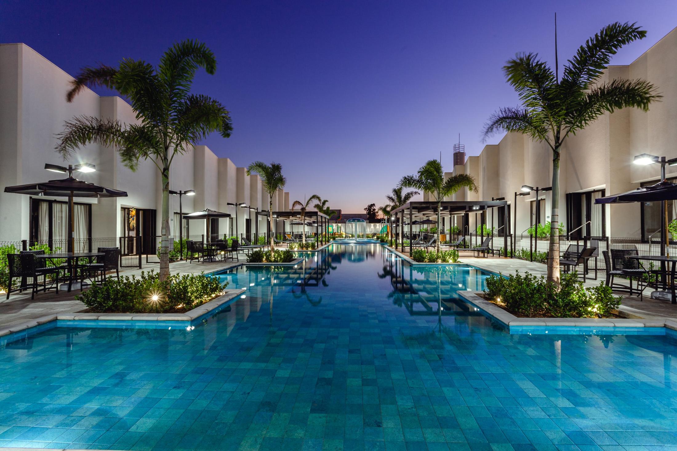 Piscina do Village Porto Rico ao entardecer, com palmeiras iluminadas e os lofts dos dois lados